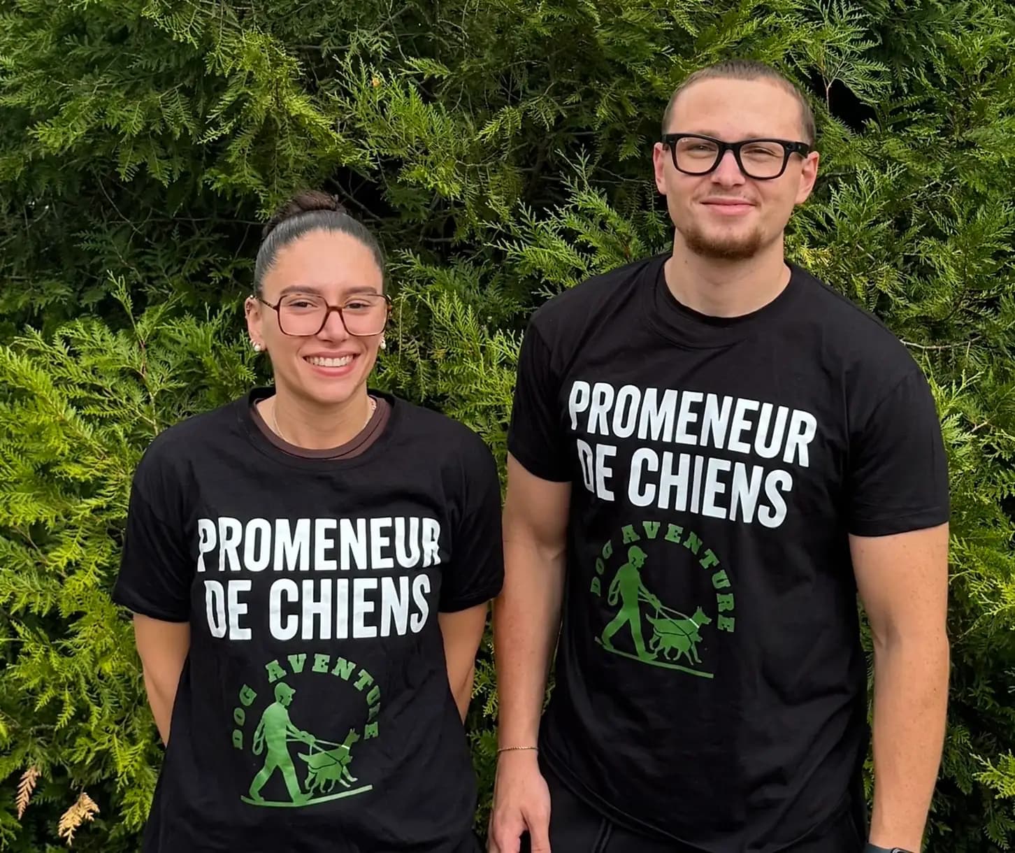 Les deux promeneurs de Dog Aventure en t-shirt « Promeneur de chiens », devant une haie
